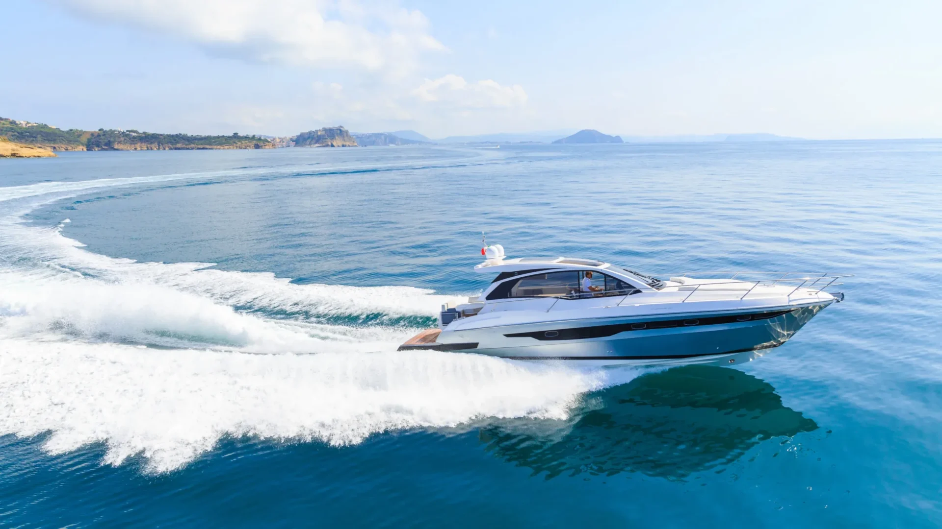 Un bateau de sport blanc et bleu à pleine vitesse au large d’une côte, sillage d’écume derrière lui