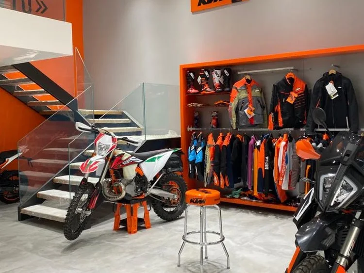 Intérieur du magasin : une enduro blanche sur béquille devant l’escalier, et un mur d’équipement orange garni de bottes, vestes et combinaisons