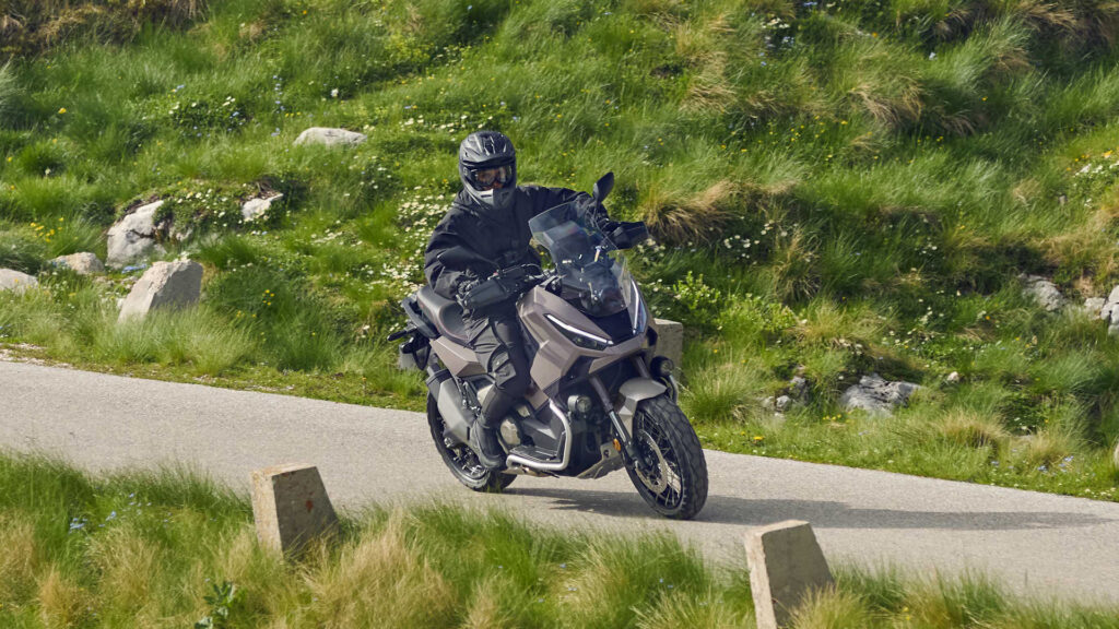 Honda X-ADV 750 en virage sur une petite route de montagne bordée d'herbe