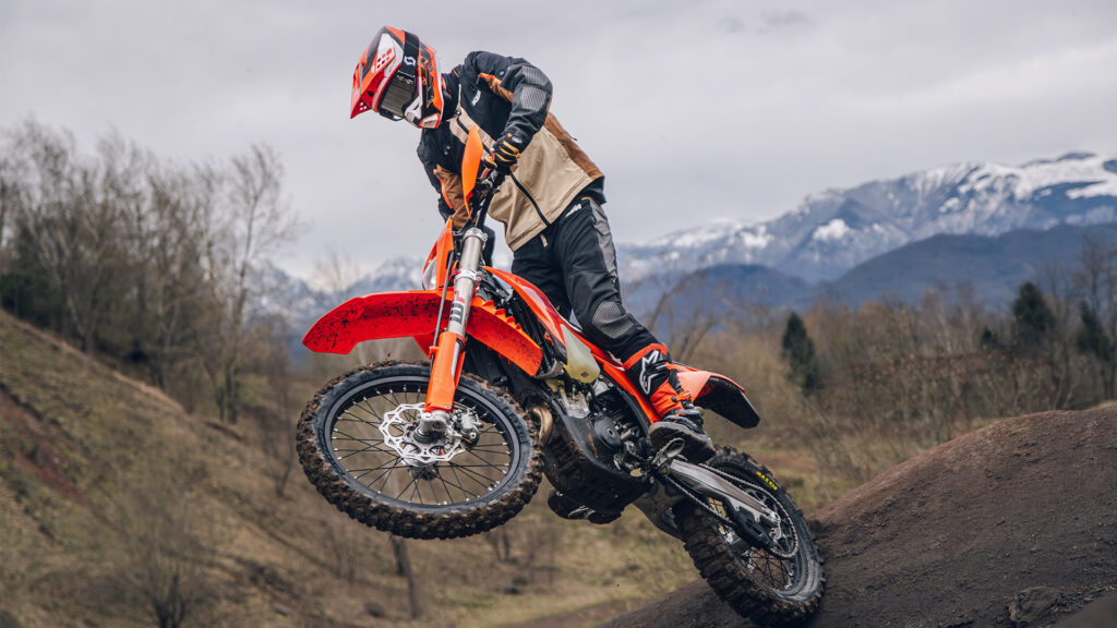 KTM 250 EXC-F 2027 franchissant une bosse de terre, montagnes enneigées en arrière-plan