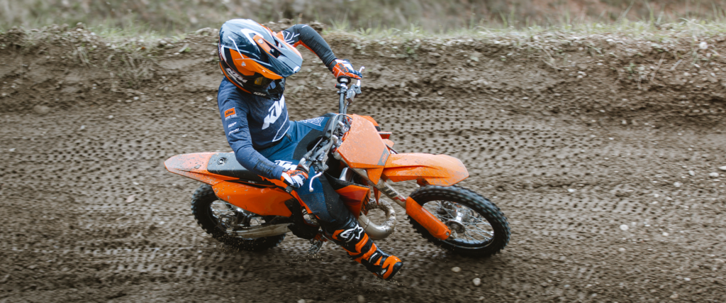 Jeune pilote casqué en tenue de cross bleue et orange sur une KTM 65 SX, en virage sur une piste de terre