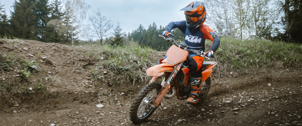 Jeune pilote casqué sur une KTM 65 SX boueuse, en appui dans un virage relevé de motocross