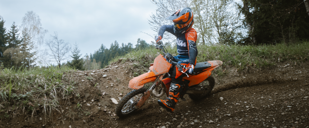 Jeune pilote casqué sur une KTM 65 SX, en sortie de virage au pied d'un talus herbeux