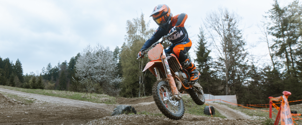 Jeune pilote casqué sur une KTM 65 SX, roue avant levée sur une bosse de piste de motocross en forêt