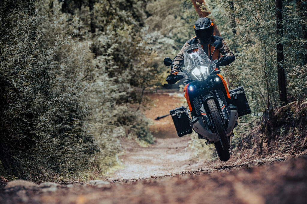 KTM 890 Adventure orange équipée de valises, pilote casqué arrivant de face sur une piste en sous-bois