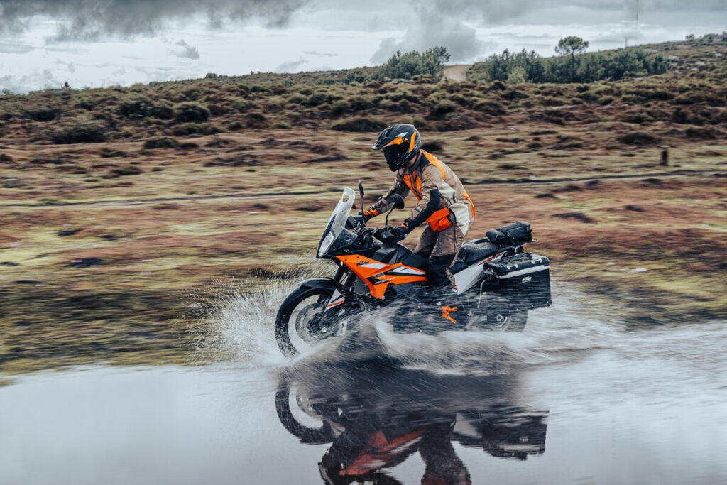 KTM 890 Adventure orange équipée de valises, pilote casqué traversant une flaque sur un plateau de lande