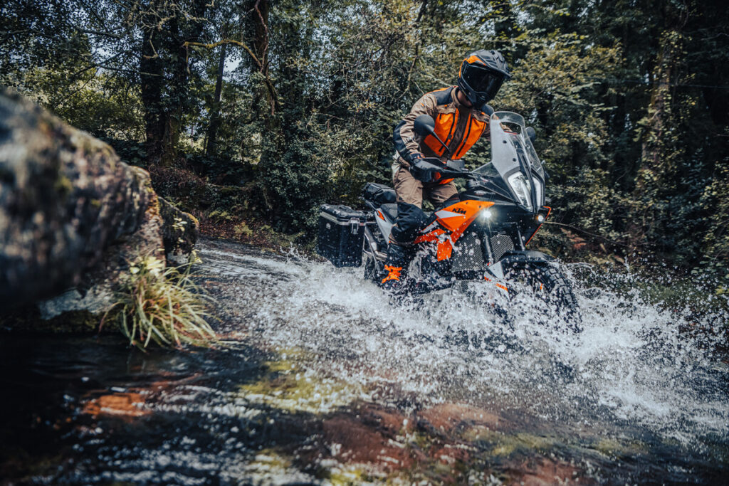KTM 890 Adventure orange équipée de valises, pilote casqué franchissant un gué dans une forêt