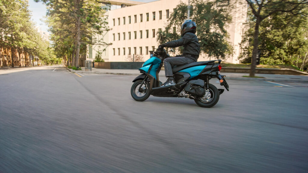 Yamaha Cygnus Ray ZR 125 cyan mat abordant un virage sur une place arborée
