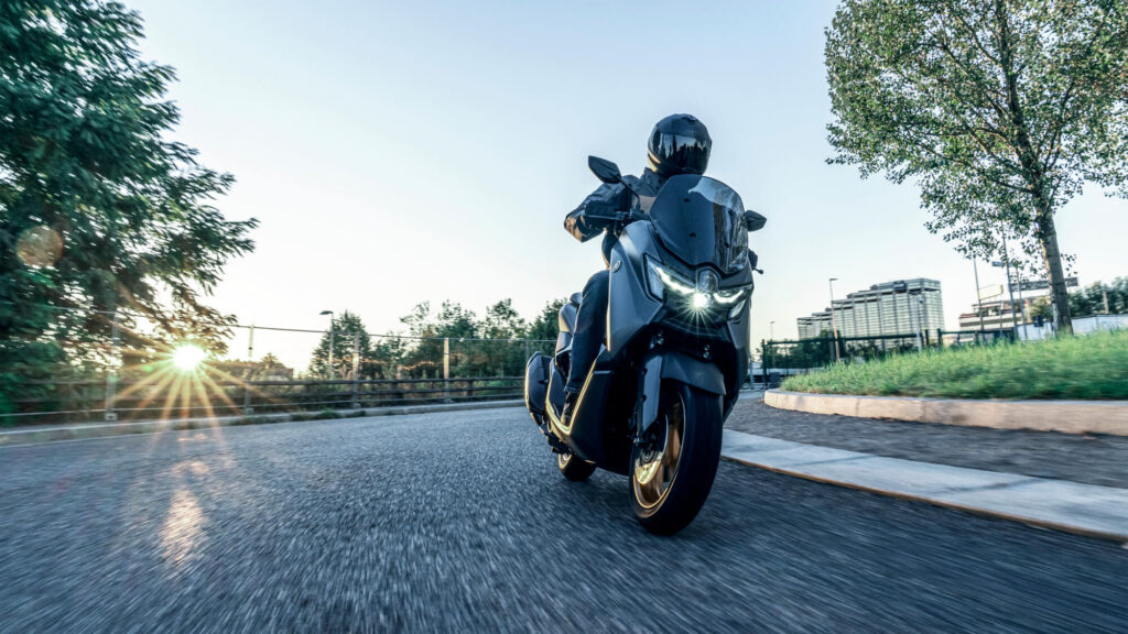 Pilote casqué sur un Yamaha NMAX Tech Max gris anthracite à jantes bronze, en roulant à contre-jour