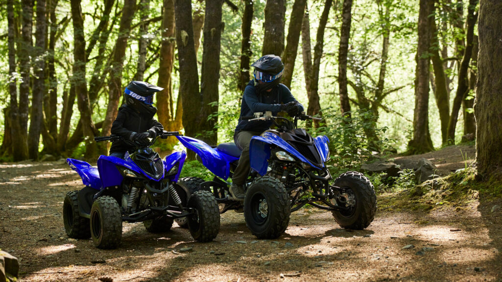Deux jeunes pilotes casqués sur des Yamaha Raptor 110 bleus en sous-bois
